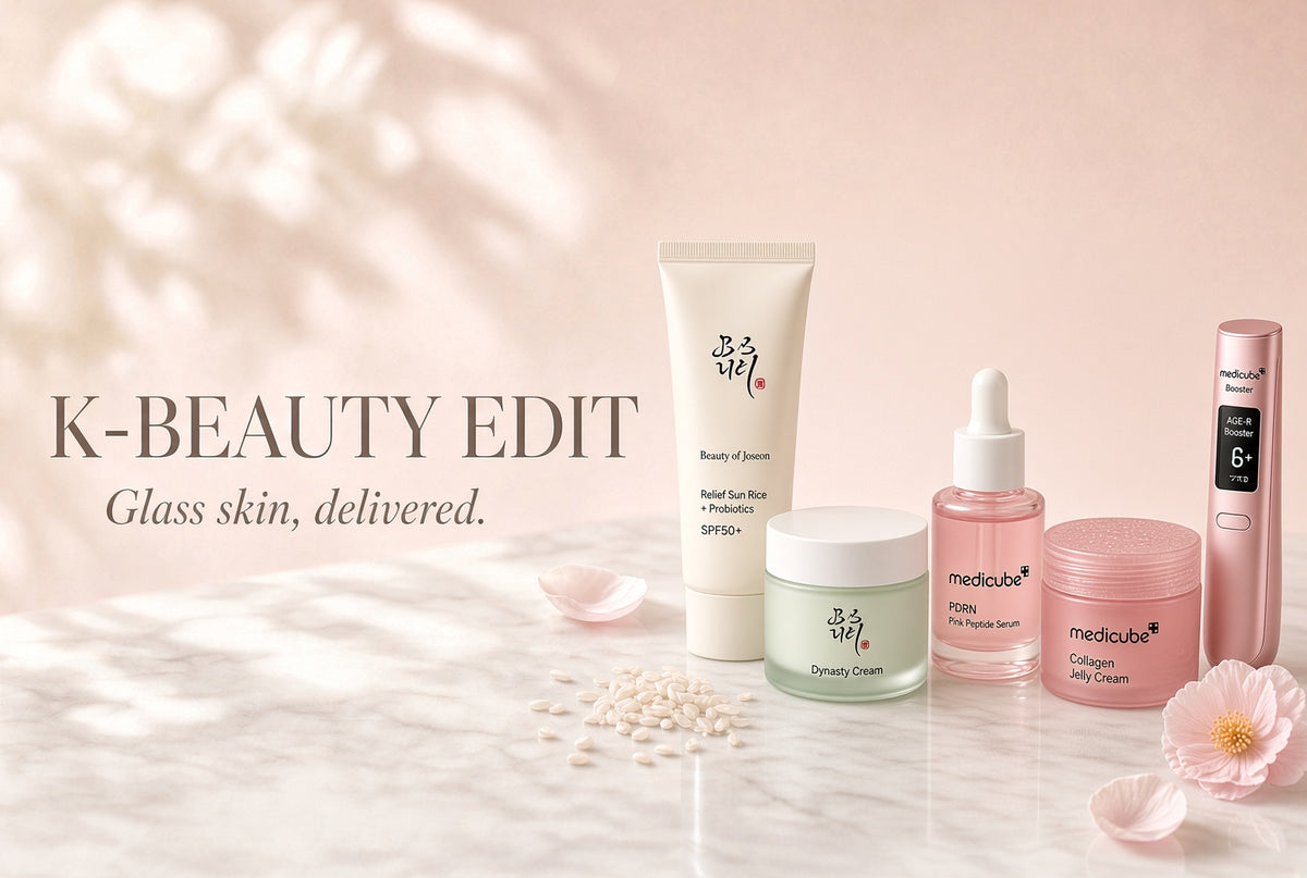 A group of K-beauty skincare products arranged on a light marble surface with soft pink shadows. Visible products include a white tube, a pale green jar, a pink dropper bottle, a pink jar, and a tall pink device.